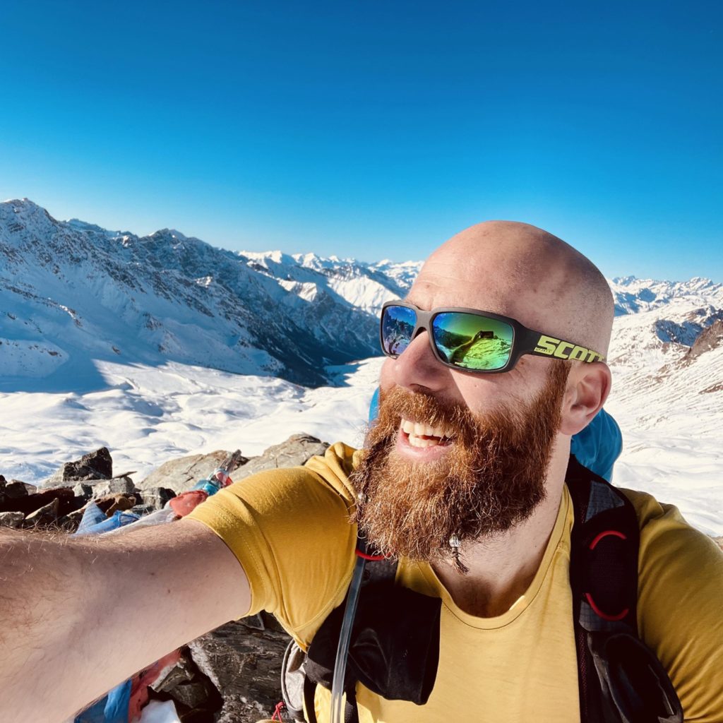 Pierre, lunettes de soleils, grosse barbe et crâne rasé, pose en souriant dans un éclatant t-shirt jaune et sac au dos au milieu de montagnes enneigées.